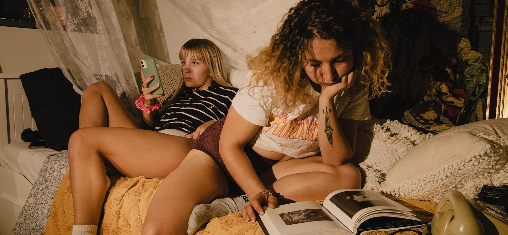 Two young women sitting on bed in underwear, one reading a book, the other on her phone