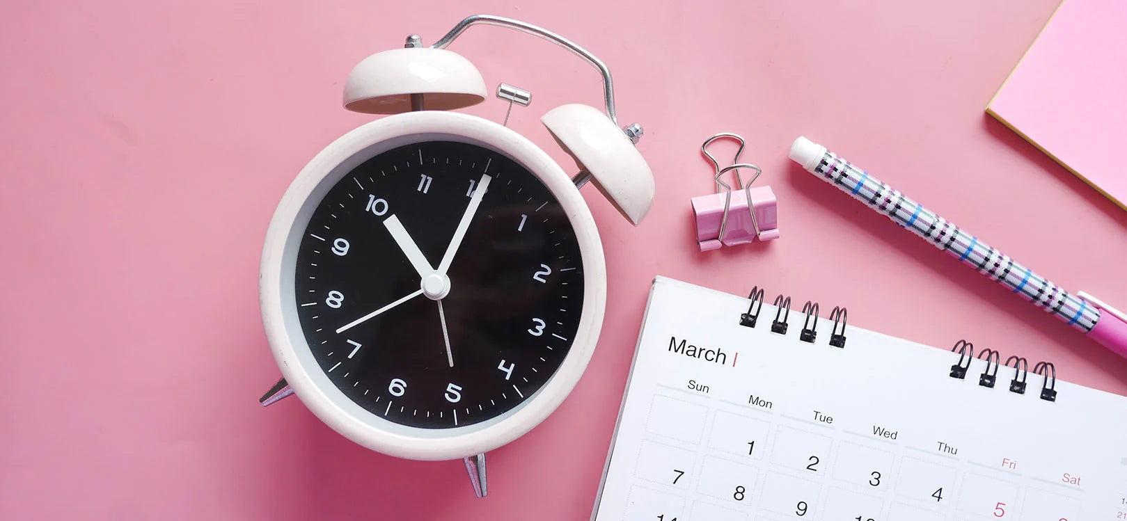 Alarm clock next to calendar & pen on pink background