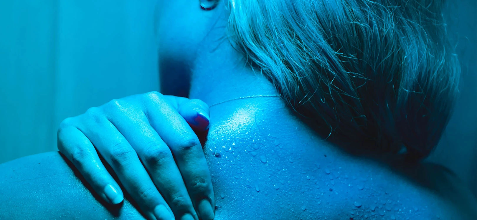 Blue-toned image of woman with blonde hair over her right shoulder holding her left shoulder and lower neck area in pain